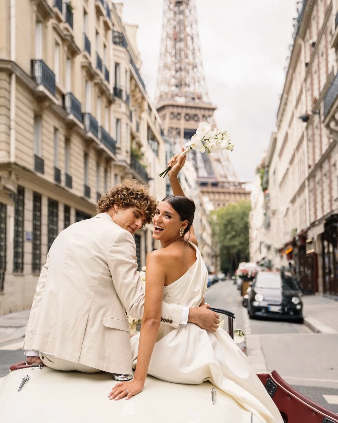 A happy couple, dressed in wedding attire, sitting on the hood of a vintage car on a city street with the Eiffel Tower in the background. The woman is holding a bouquet of white flowers and smiling at the camera, while the man looks over his shoulder.