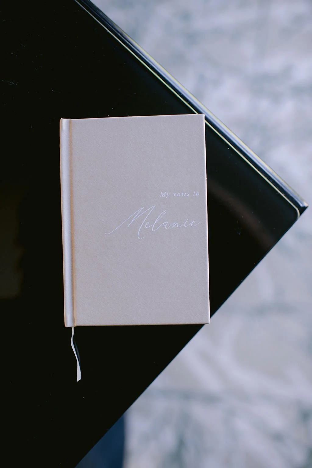 A white hardcover book with the title 'My vows to Melanie' written in elegant cursive is resting on a black surface, with a black electronic device partially visible underneath it.