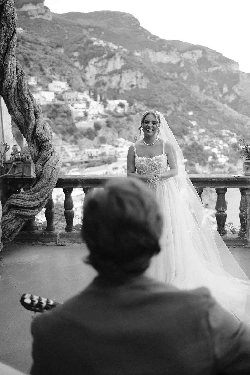 A bride in a wedding dress smiling at someone, possibly her groom, on a balcony with a mountain landscape in the background. A person in the foreground appears to be holding a guitar.