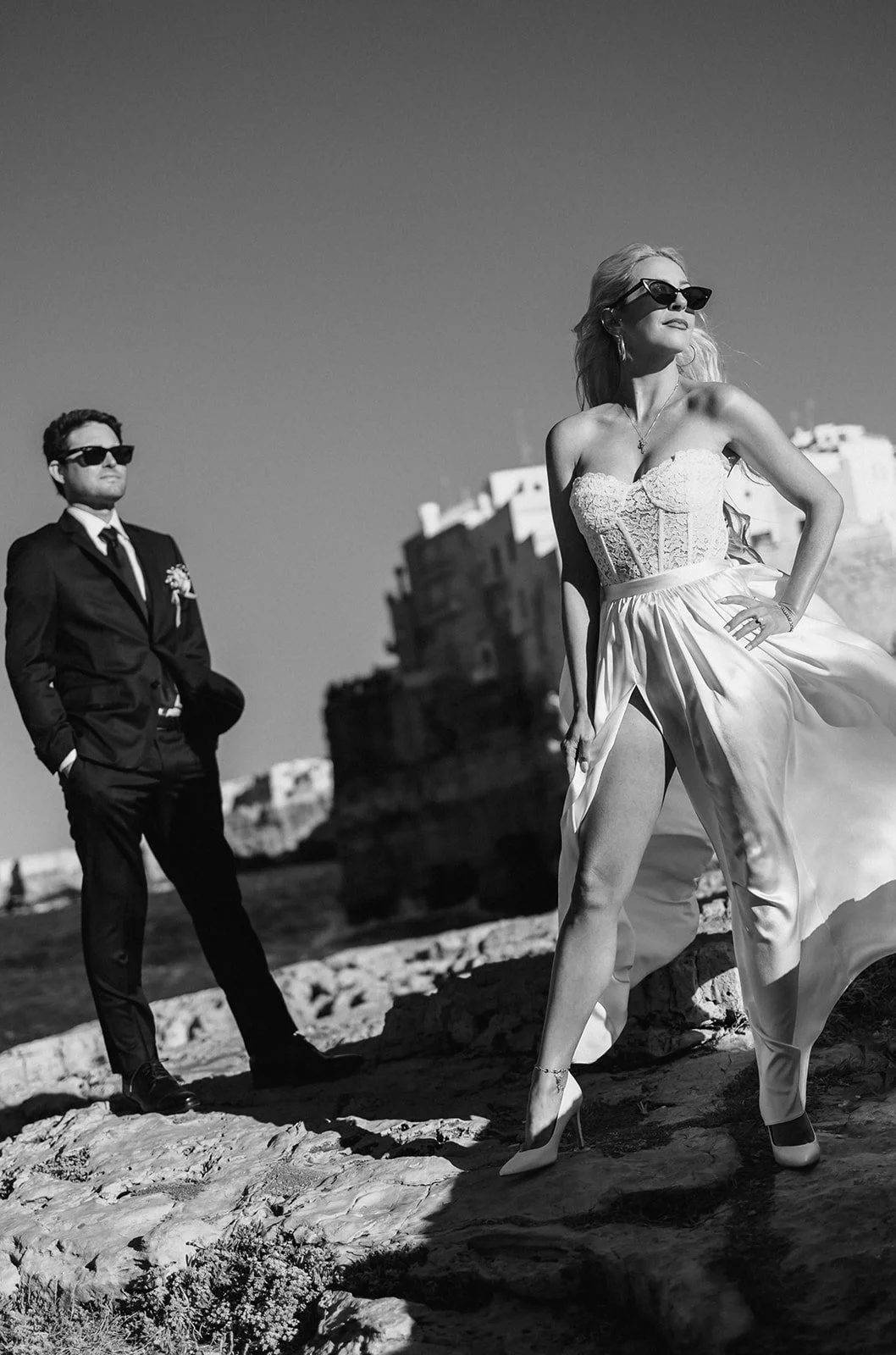 A black and white photo of a woman in a strapless dress with slit pants posing confidently with her hand on her hip, and a man in a suit with sunglasses standing in the background. They are outdoors with old buildings and rocky terrain visible.