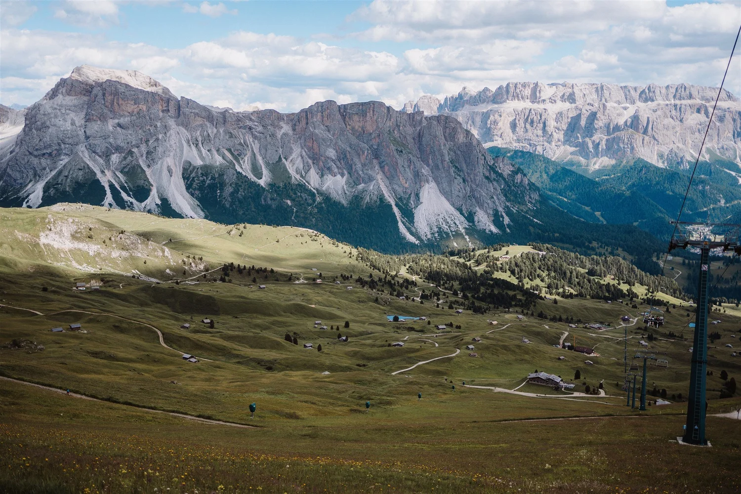 Mountain landscape with rocky cliffs behind a green valley dotted with small houses and winding roads, with a chairlift cable visible on the right side.
