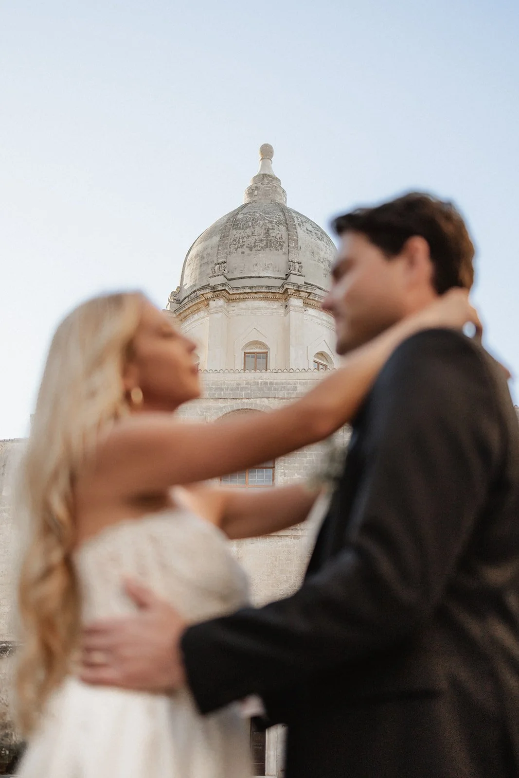 A couple dancing close together in front of a historic building with a domed roof, with the woman’s arm around the man's neck and the man holding her waist.