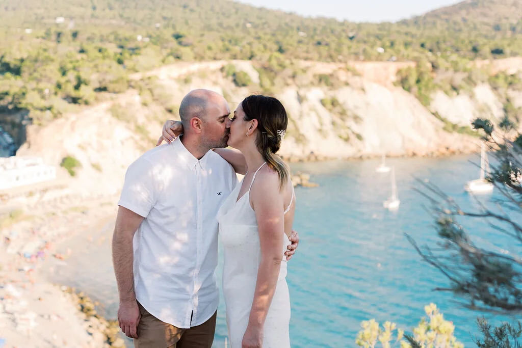 A couple sharing a kiss near a scenic coastal area with boats on the water and hillside in the background.
