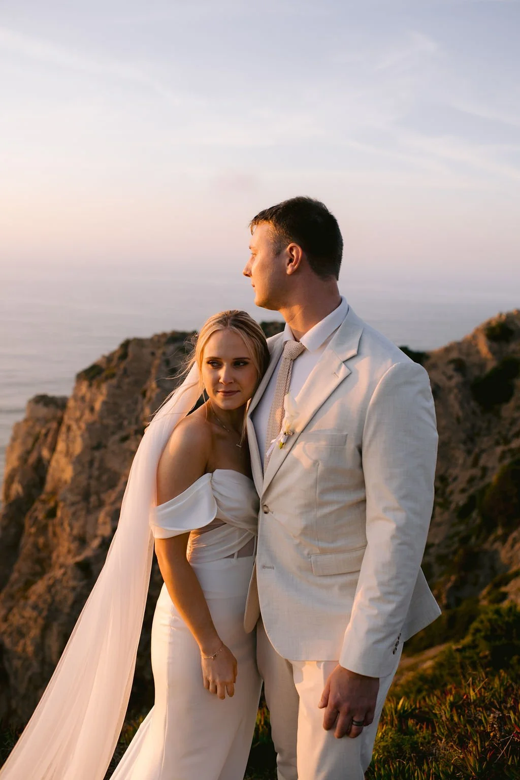 A bride and groom in wedding attire standing on a cliffside during sunset, with a view of the ocean and distant cliffs in the background.