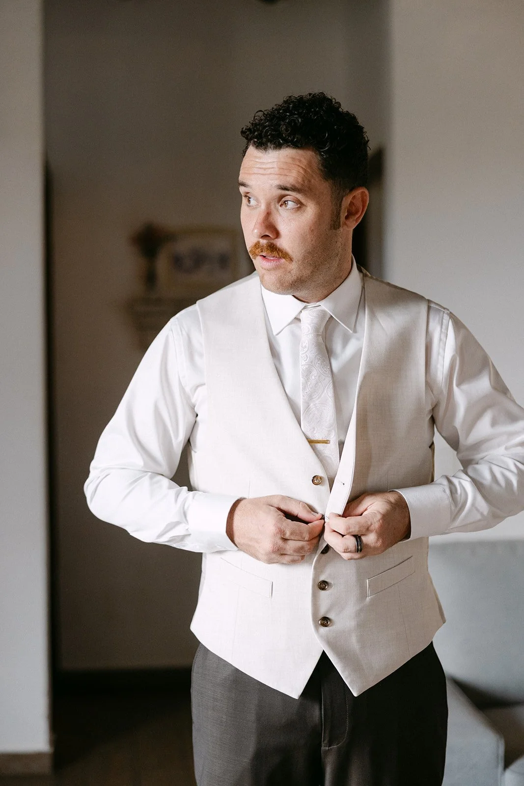 Man in white vest and shirt buttoning his vest, looking to the side.