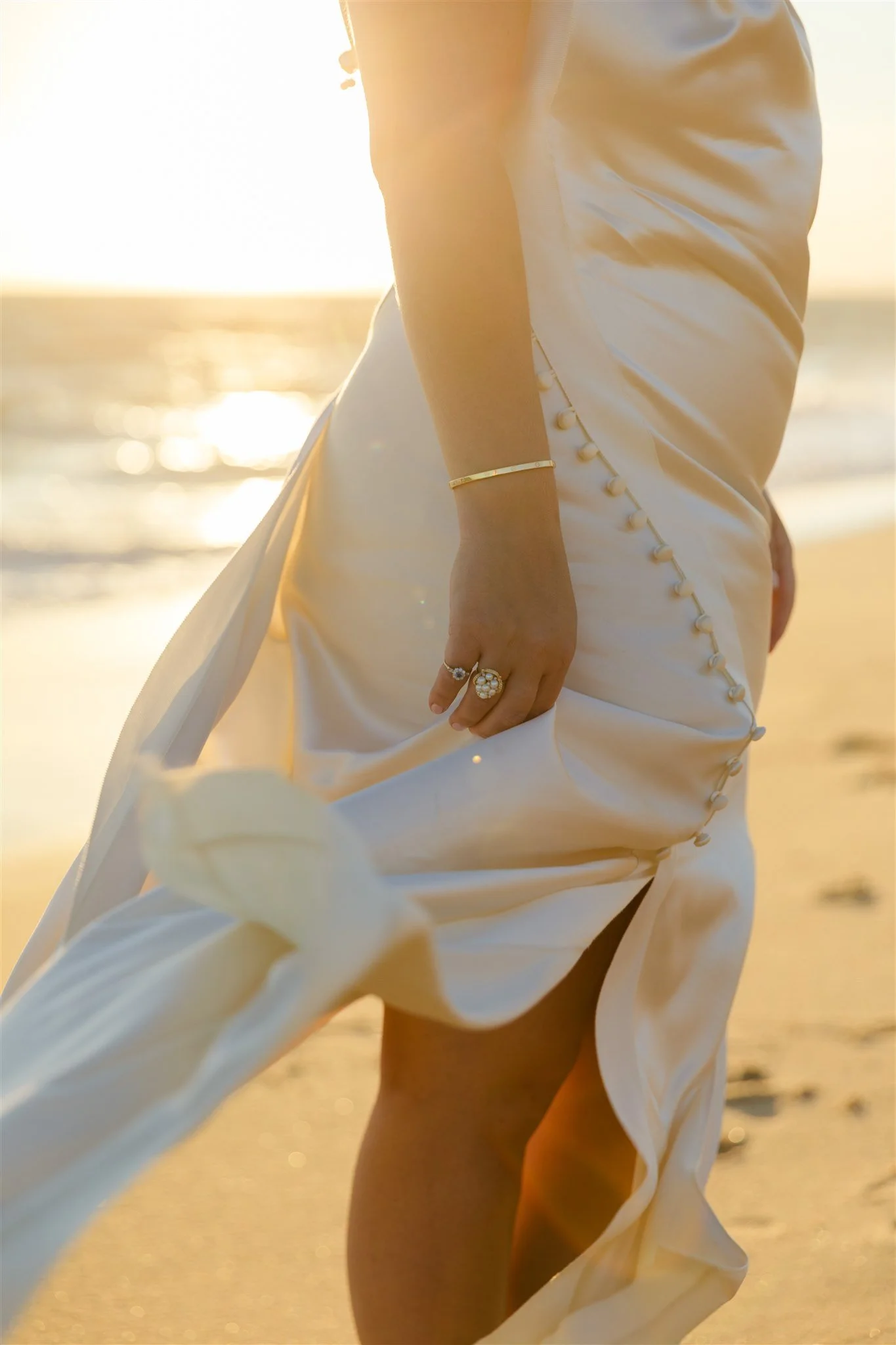 Close-up of a woman's lower body in a white satin dress with buttons on the side and rolled-up sleeves, standing on a sandy beach at sunset.