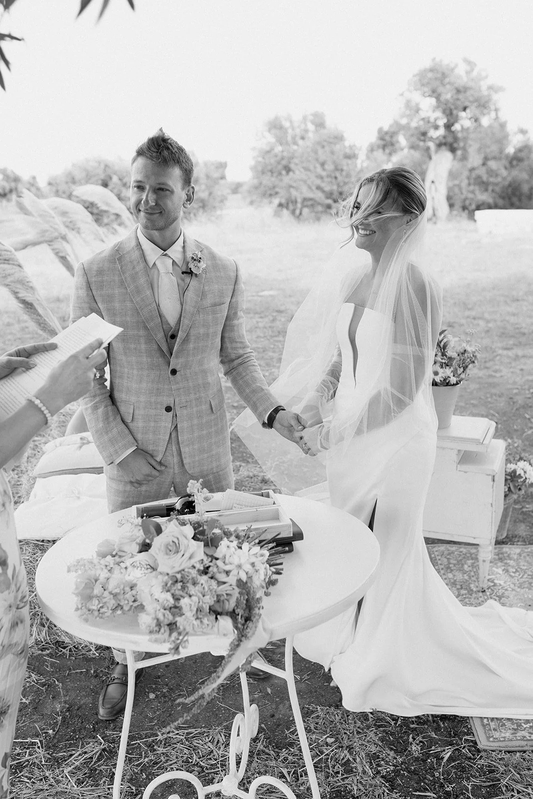 A black-and-white photo of a wedding ceremony outdoors showing a groom and bride holding hands, with a person reading from a book. The groom is in a light-colored checkered suit, and the bride in a white dress with a veil. There is a small round tabl