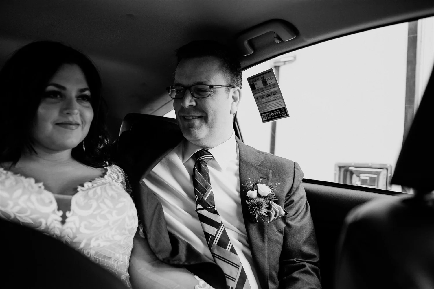 A black-and-white photo of a smiling couple seated in an airplane. The woman is wearing a dress with lace details, and the man is dressed in a suit with a tie and glasses. They are looking at a device with a boarding pass or ticket visible nearby.