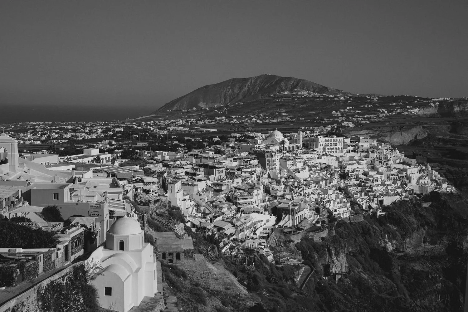 A black and white aerial view of a hilltop town with numerous white buildings, a church in the foreground, and mountains in the background.