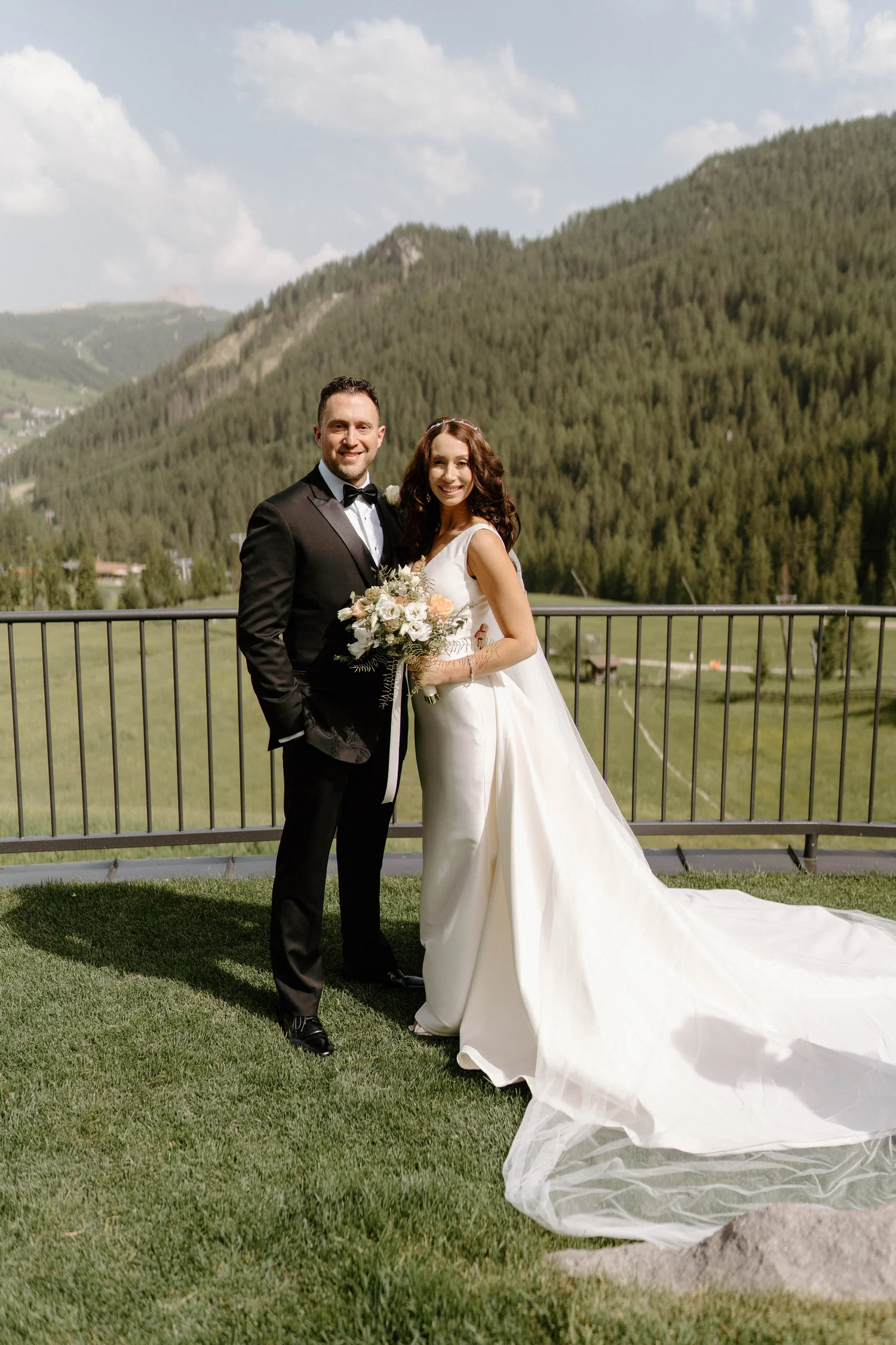A bride and groom stand outside on a grassy area with mountains and trees in the background. The groom is wearing a tuxedo, and the bride is in a white wedding dress holding a bouquet of flowers.