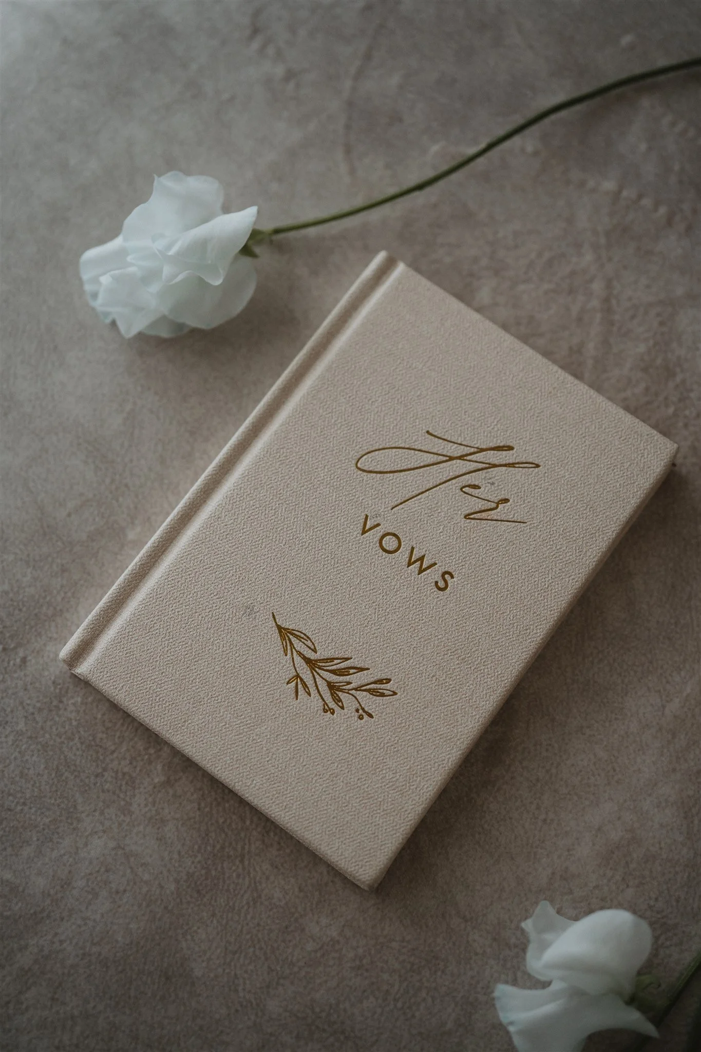 A closed beige book with the title in gold lettering and a decorative leaf design on the cover, with a white flower with a long green stem lying next to it on a textured surface.