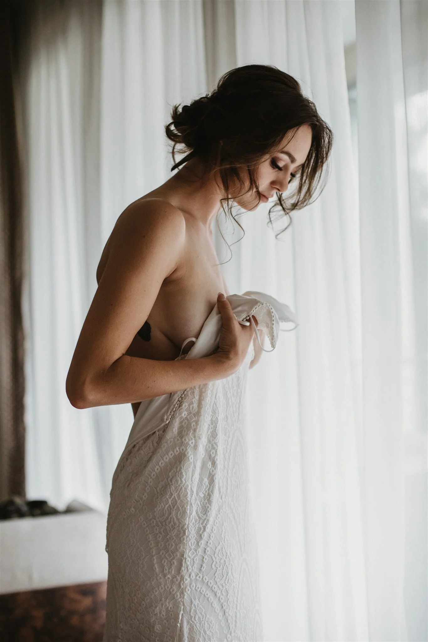 A woman with brown hair styled in loose curls, wearing a white lace dress, standing in front of white curtains, looking down and holding the dress with one hand.