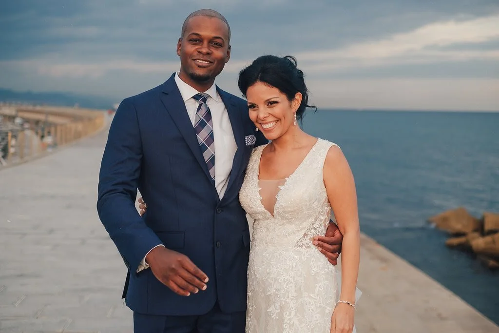 A smiling couple dressed in wedding attire stands on a pier by the ocean, with a cloudy sky in the background.