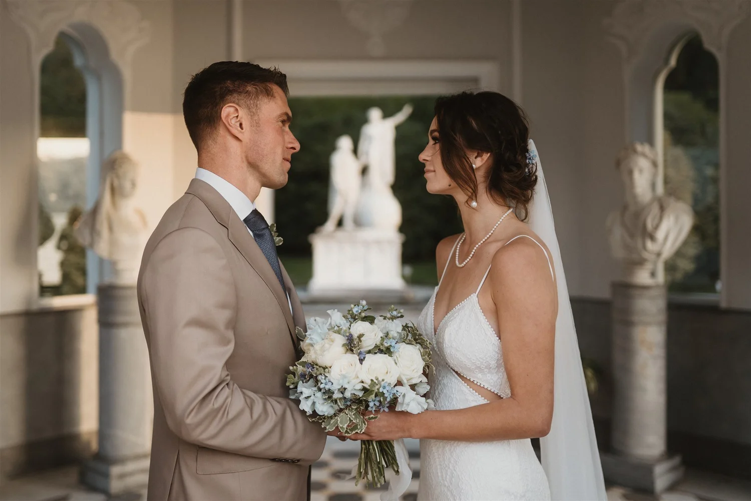 A bride and groom face each other during a wedding ceremony, holding a bouquet of white and blue flowers, in an outdoor space with classical statues in the background.