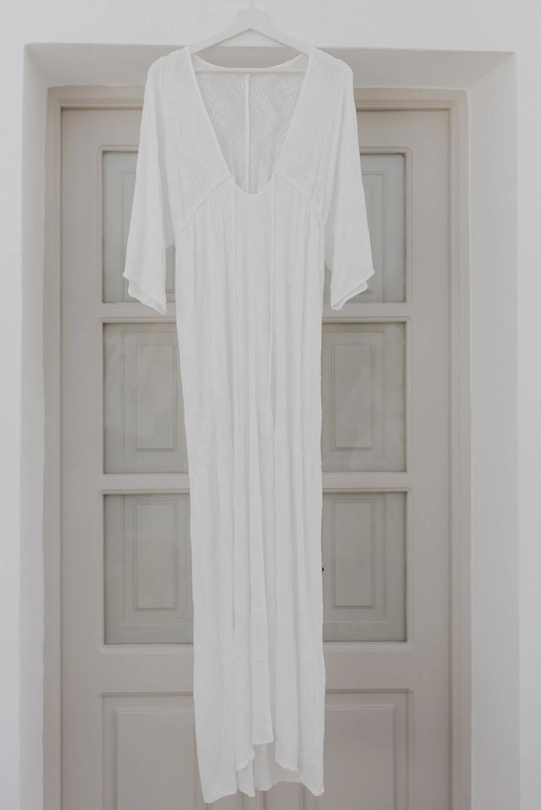 A white, long, flowy dress with three-quarter sleeves hanging on a white hanger in front of a paneled door.