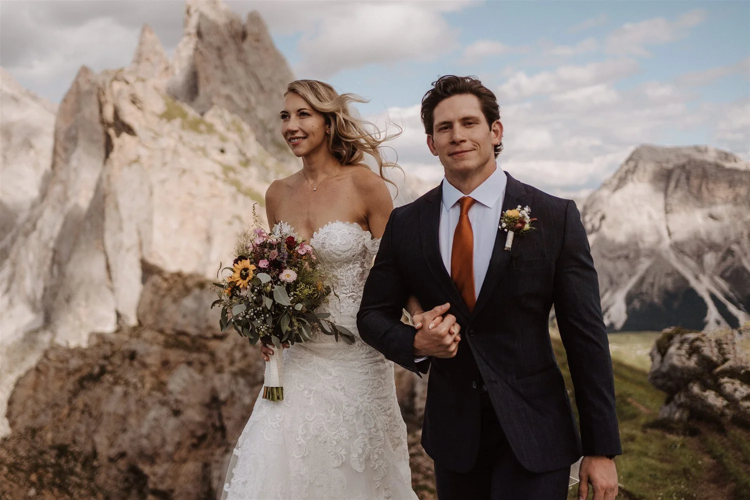 A bride and groom standing outdoors with rocky mountains in the background. The bride is wearing a white lace wedding dress and holding a bouquet of flowers. The groom is dressed in a dark suit with a white shirt and a burnt orange tie, clasping hand