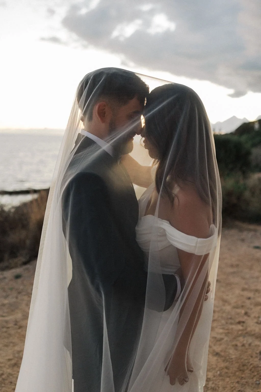 A newlywed couple standing close together on a beach during sunset, covered by a sheer veil, smiling and touching foreheads.