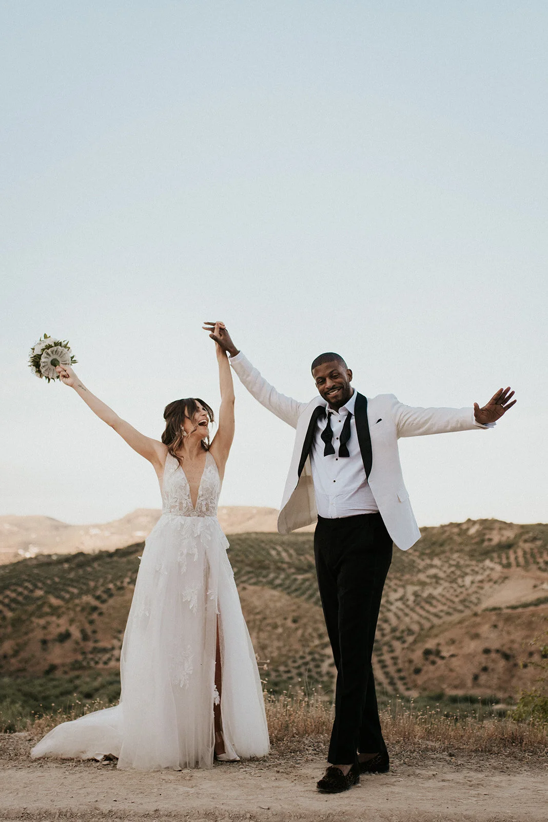A happily married couple, a woman in a white wedding dress and a man in a white suit jacket and black pants, celebrating outdoors with a scenic mountainous background.