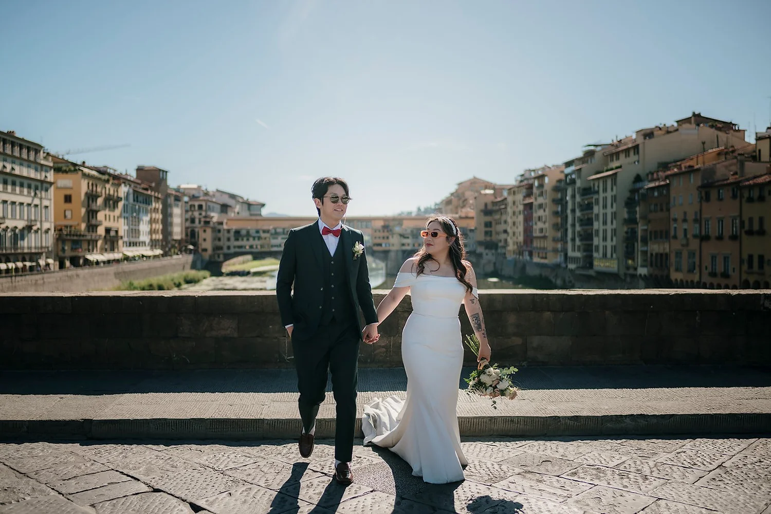 A bride and groom holding hands and walking along a waterfront promenade with city buildings in the background on a sunny day.