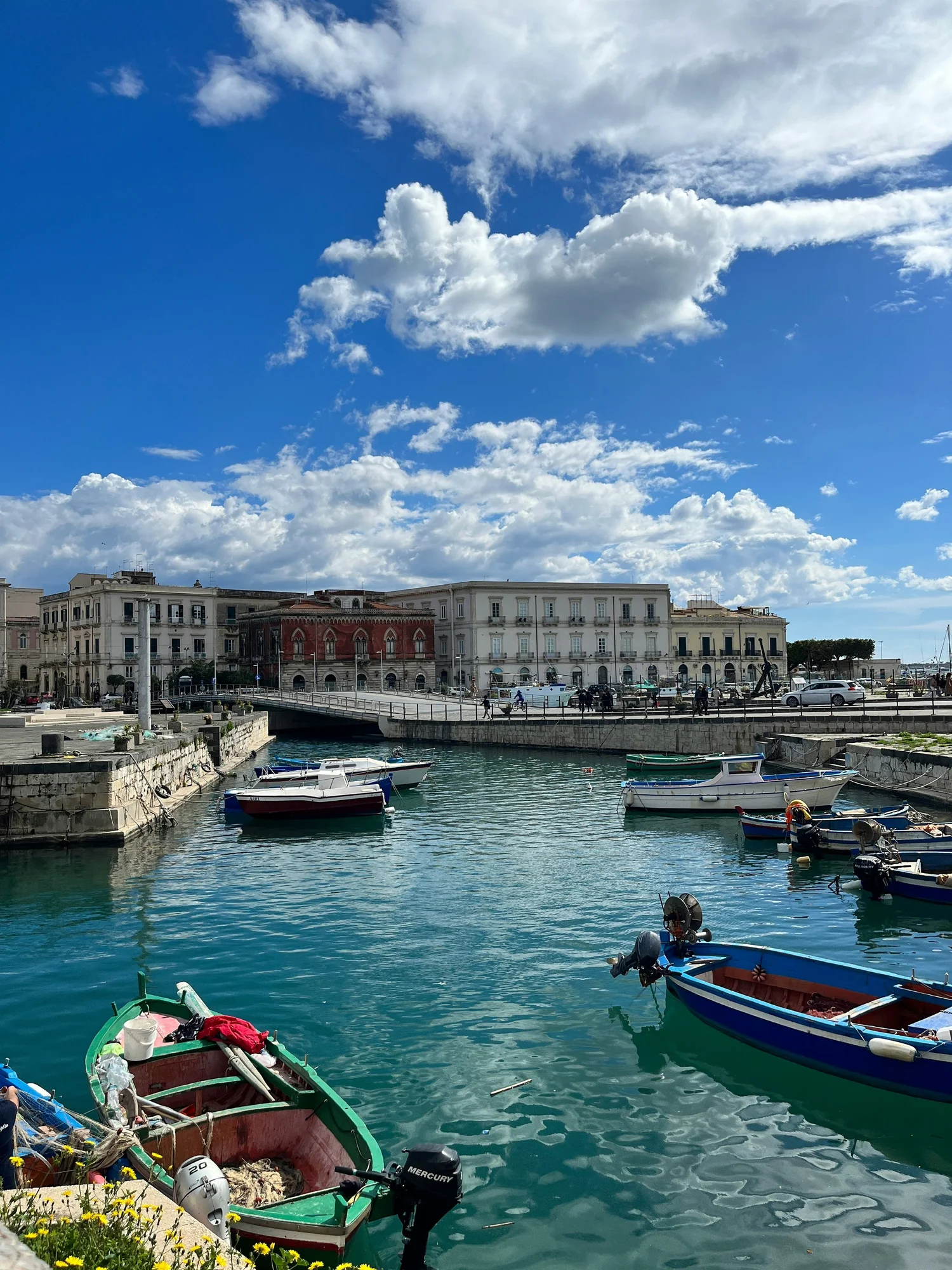A marina with small boats docked in turquoise water, with multi-story buildings and a partly cloudy sky in the background.