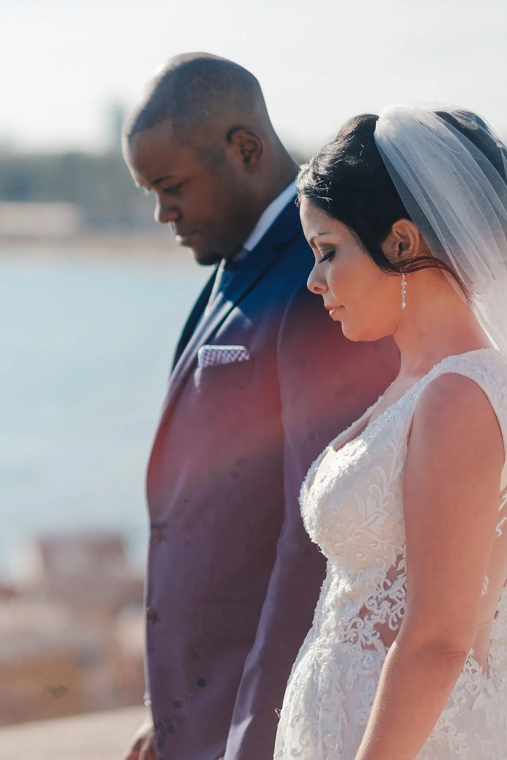 Bride and groom standing outdoors near water, dressed in wedding attire, with their heads bowed in prayer or reflection.