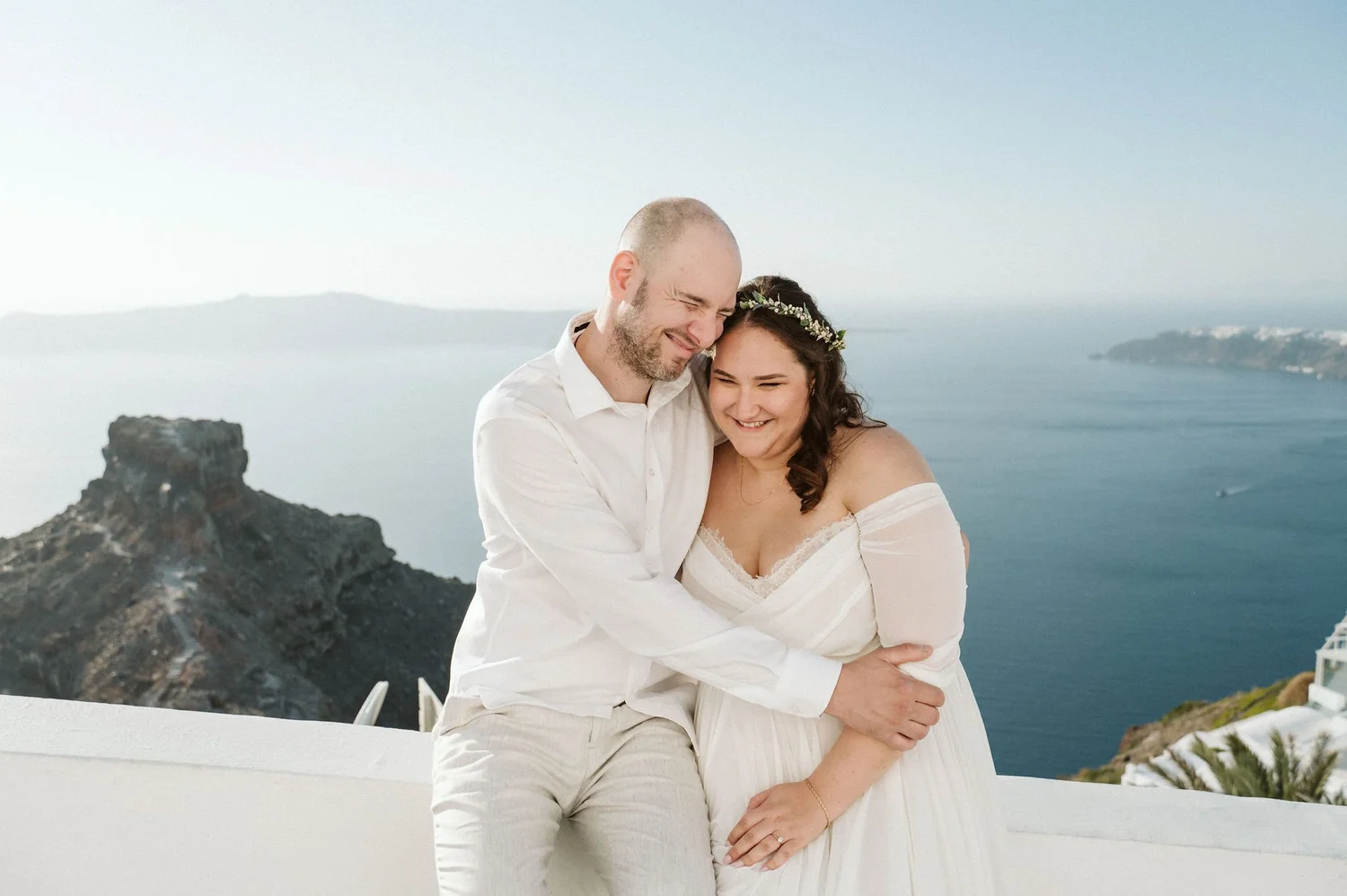 A happy couple in wedding attire embracing outdoors with a scenic ocean view and rocky cliff in the background.