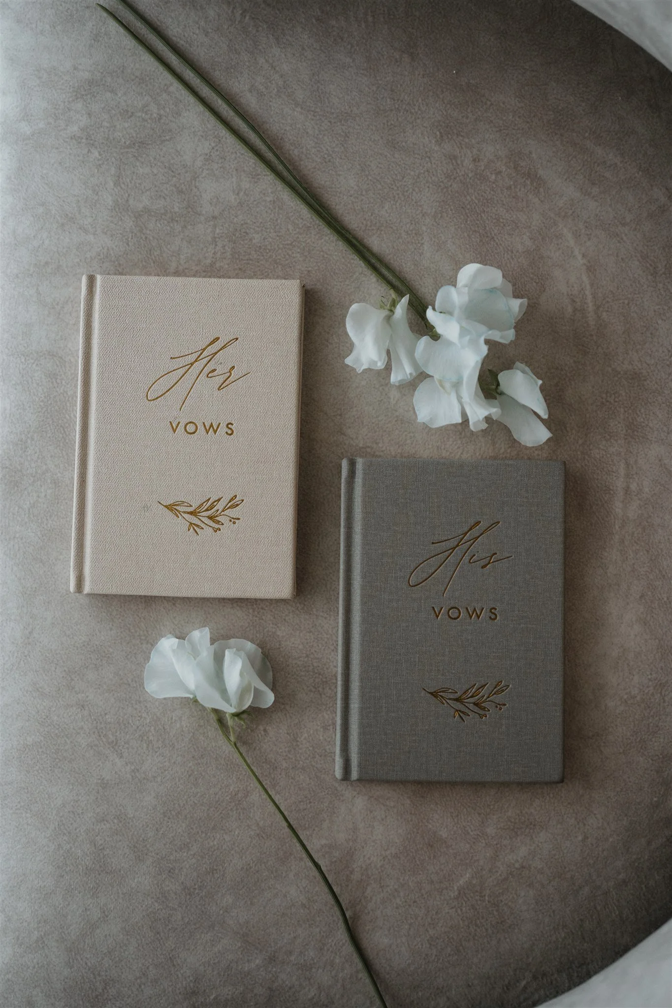Two vow books, one cream-colored and one gray, with gold lettering and decorative designs, placed on a beige surface alongside white flowers.