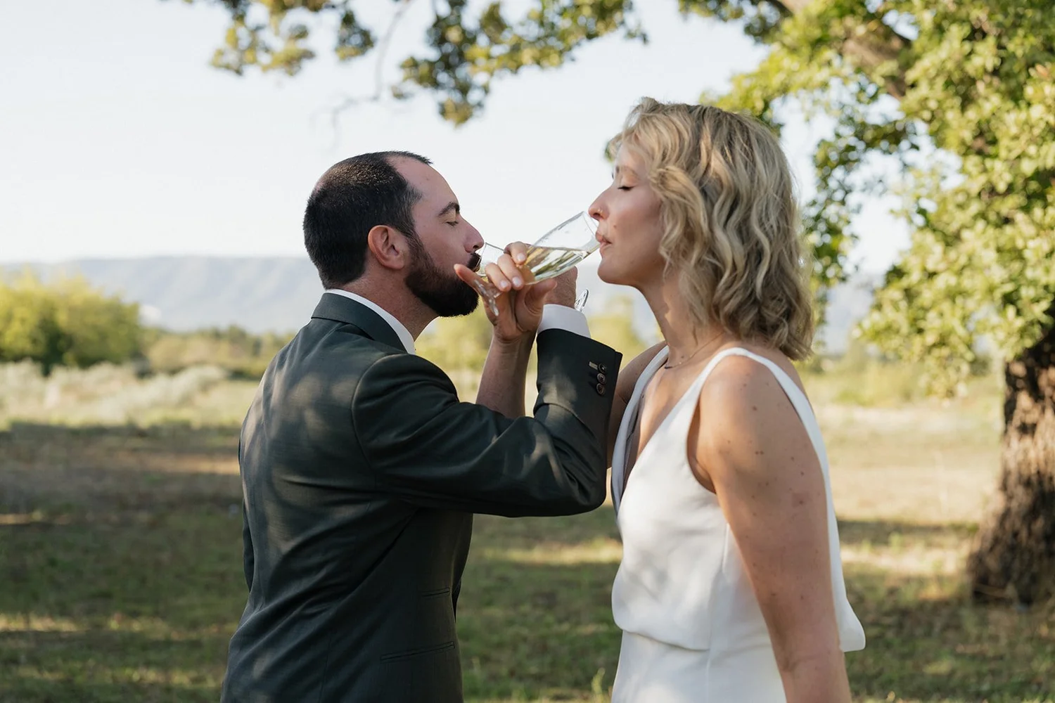 A couple at a wedding outdoors, with the groom in a tuxedo and the bride in a white dress, sharing a drink from the same champagne flute.