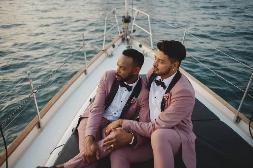Two men in pink tuxedos and black bow ties sitting on a boat at sunset, holding hands, with water in the background.