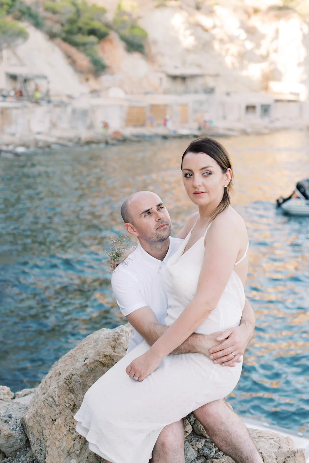 A couple sitting on rocks by the water, with a scenic background of cliffs and buildings, during sunset.