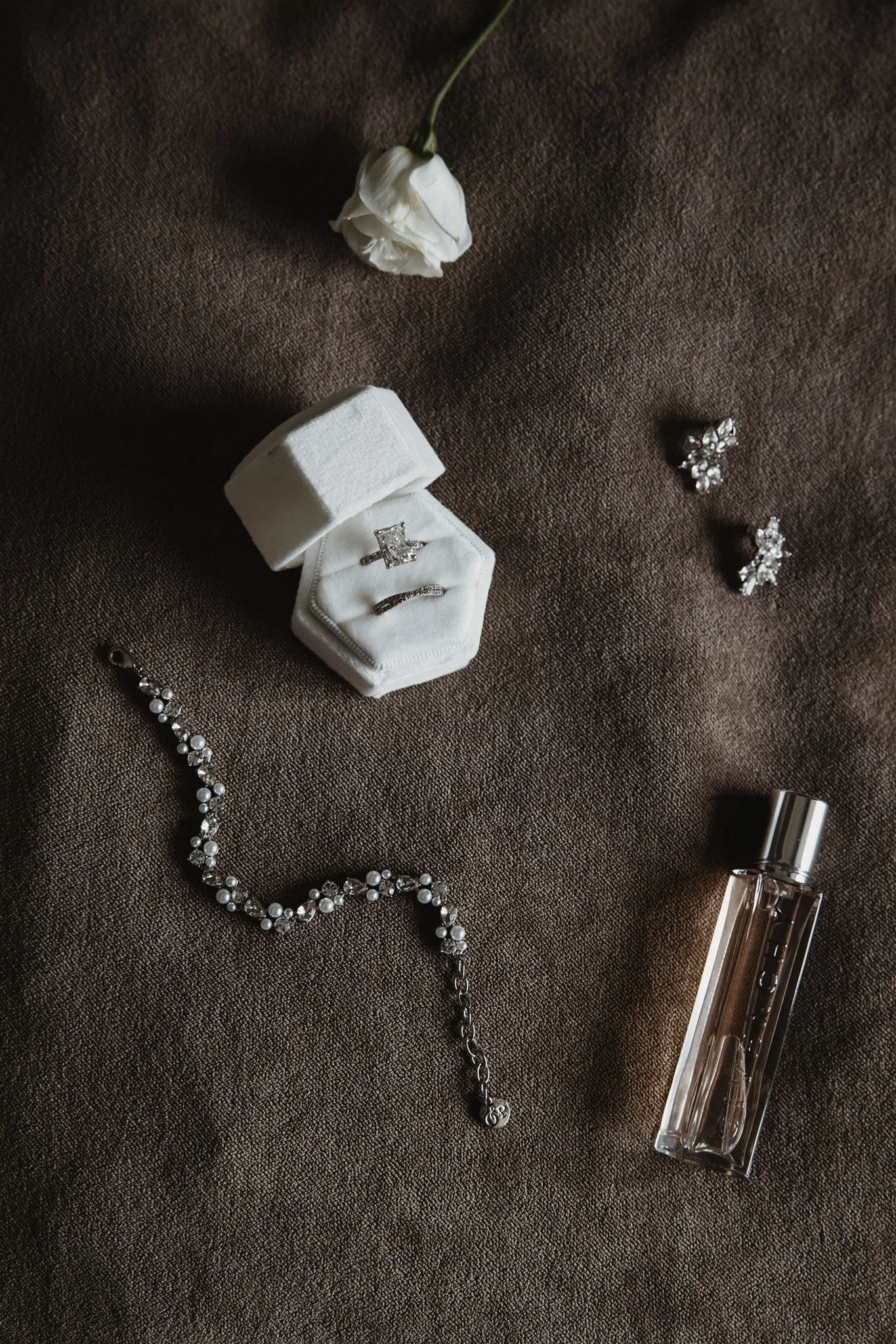 A white rose, diamond jewelry including earrings, a ring, a pearl necklace, a perfume bottle, and a cosmetic tube are displayed on a brown textured fabric background.