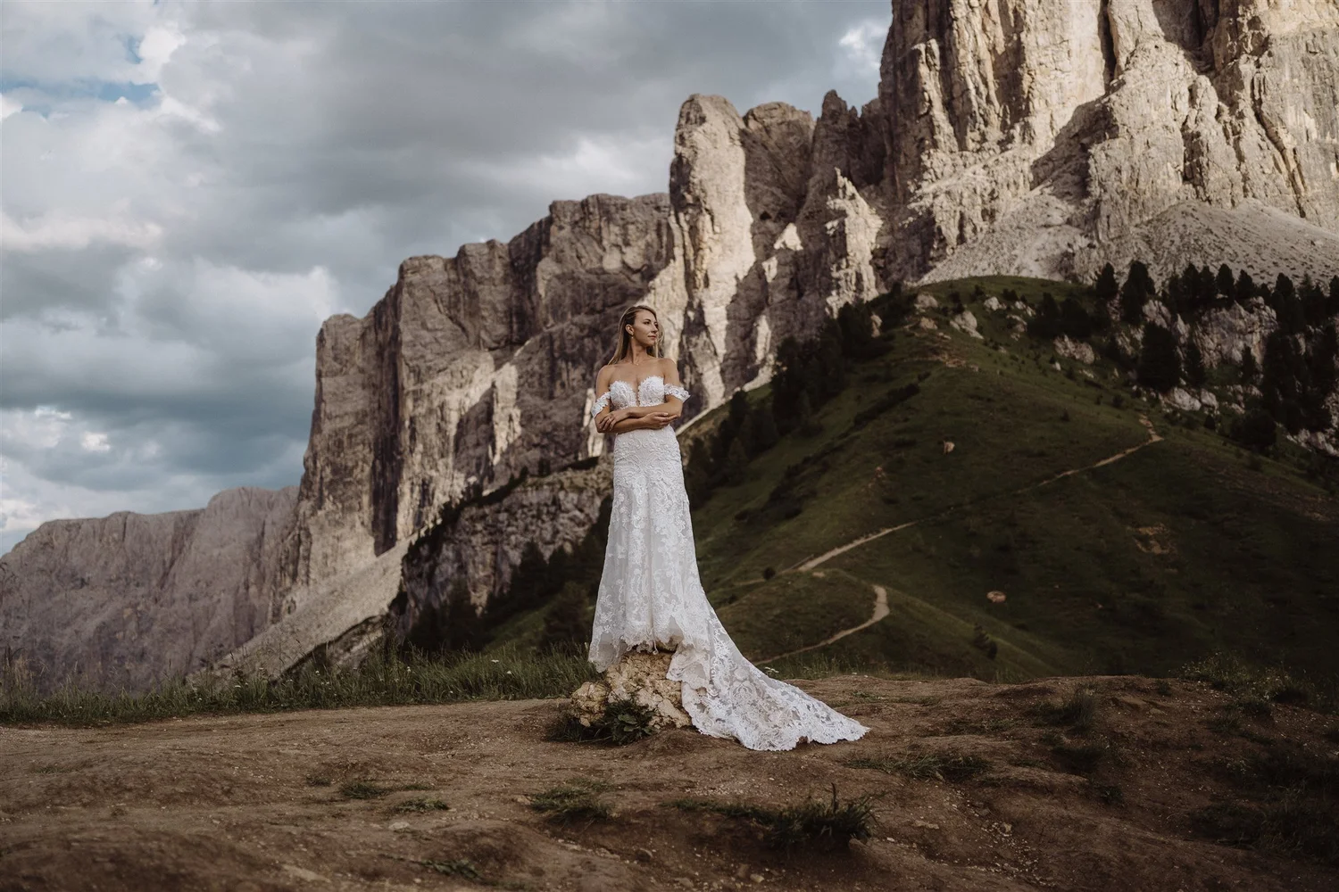 A woman in a white lace wedding dress standing on a rock in a mountainous landscape with green hills and rocky cliffs, under a cloudy sky.