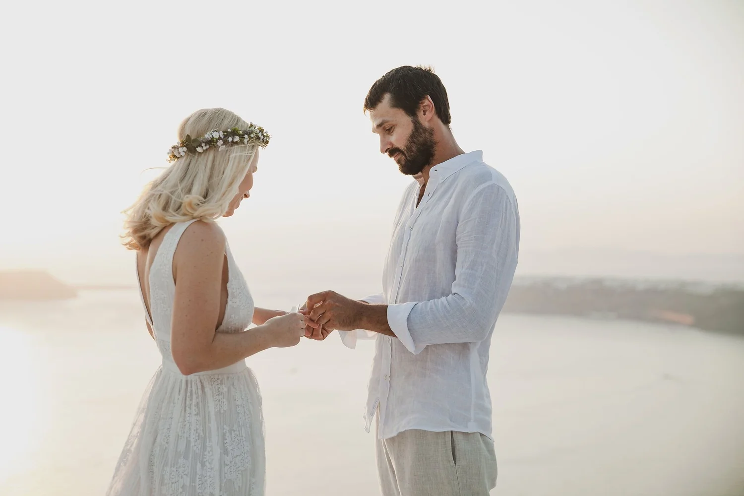 A man and woman stand close together on a beach at sunset, with the man placing a ring on the woman's finger during a wedding ceremony.
