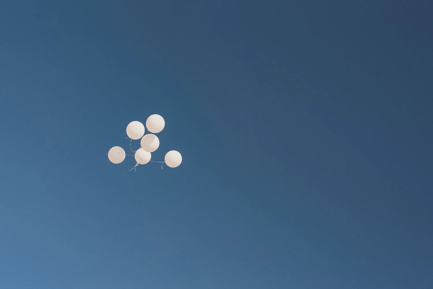 Seven white balloons floating against a clear blue sky.