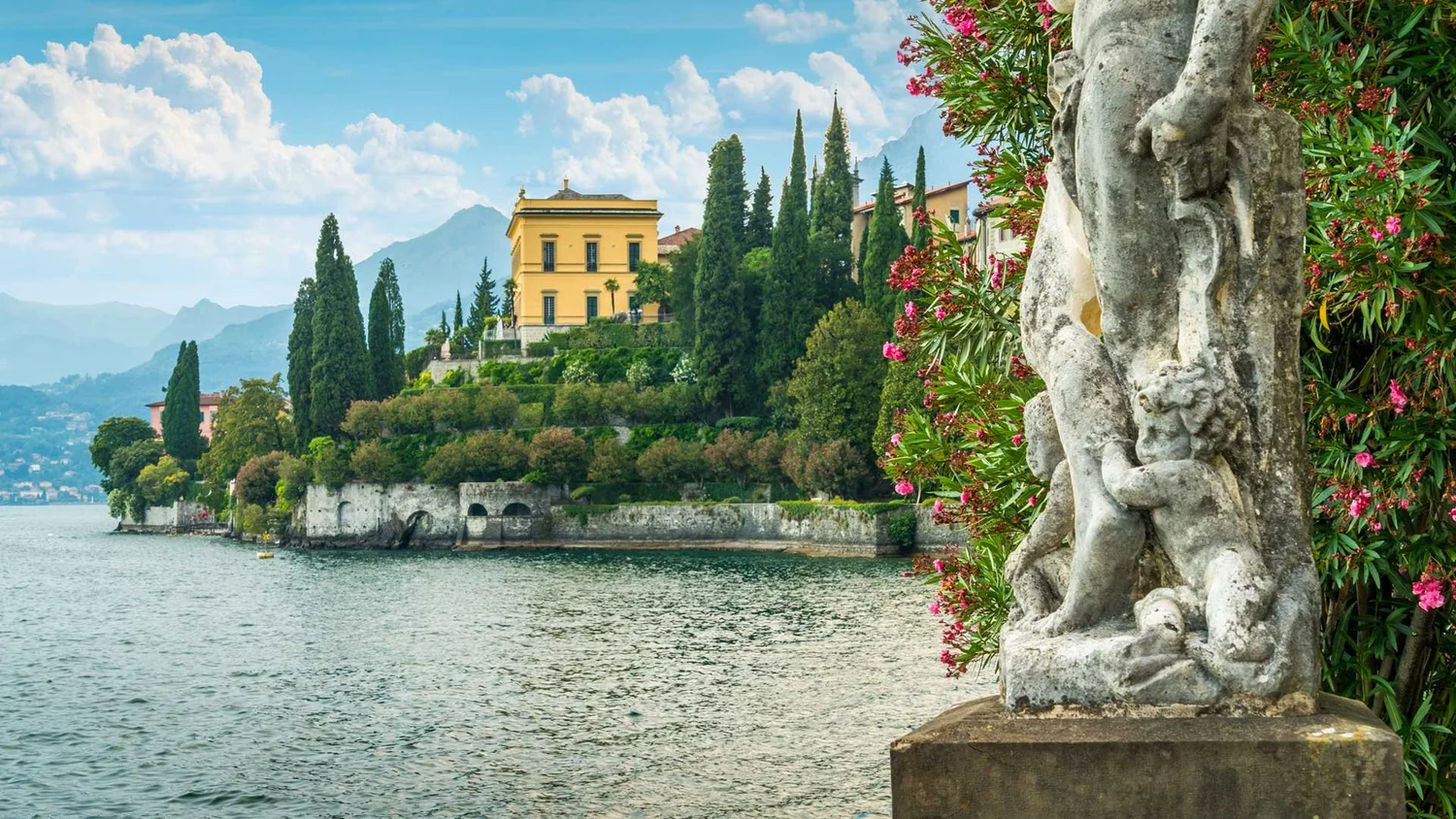 A lakeside scene with a yellow villa surrounded by tall cypress and other lush trees on a hill. The lake is calm with gentle ripples. In the foreground, there is a weathered stone sculpture of a cherub and plant with pink flowers on the right side.