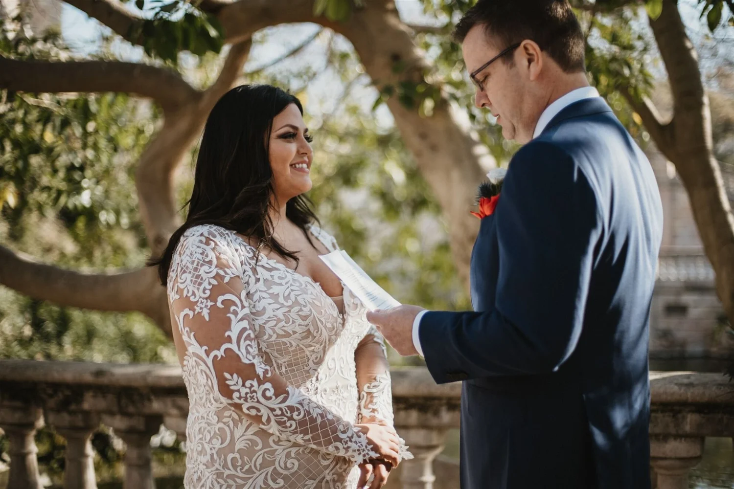 A woman in a white lace wedding dress smiling at a man in a navy suit with glasses during a wedding ceremony outdoors, with trees in the background.