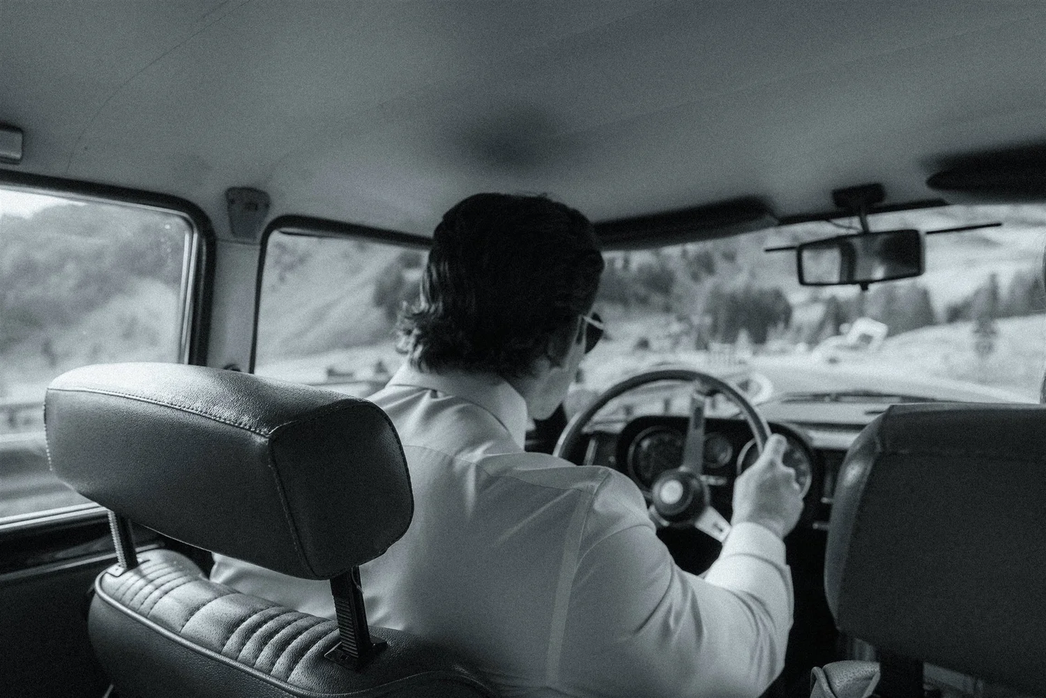 A man with dark hair and a white shirt driving a vehicle, viewed from behind inside the car. The vehicle appears to be a bus or van, with a view of the road and snow-covered landscape outside.
