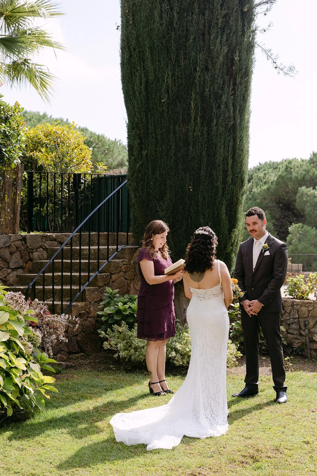 A bride and groom exchanging vows during an outdoor wedding ceremony, with a officiant looking on, in a lush garden setting.