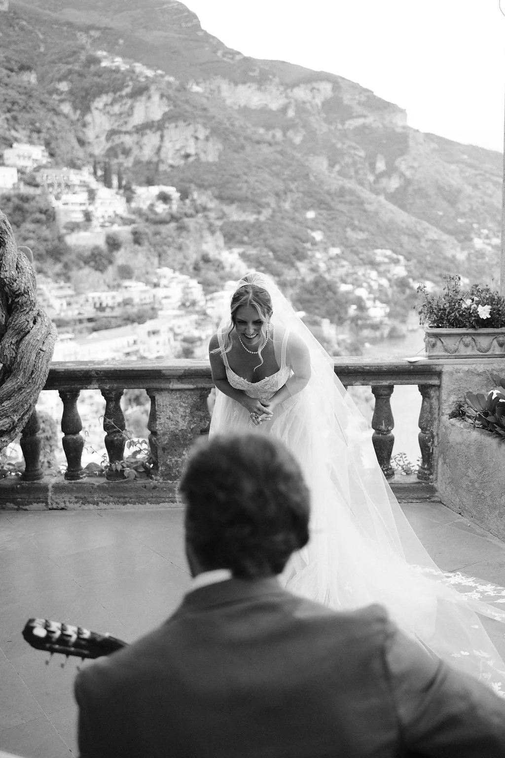 A bride in a wedding dress and veil smiling and bowing toward a groom playing guitar during a wedding ceremony outdoors with scenic mountainous landscape in the background.