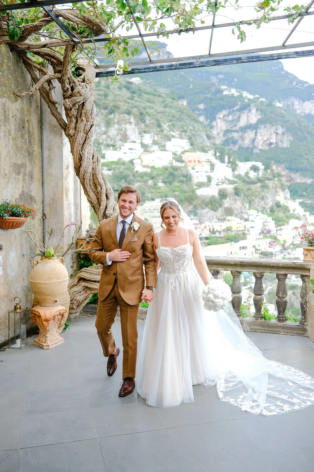 A newlywed couple walking hand in hand on a terrace with scenic hillside view in the background. The groom is wearing a brown suit with a white rose boutonniere. The bride is in a white wedding gown holding a bouquet of white roses. They are smiling