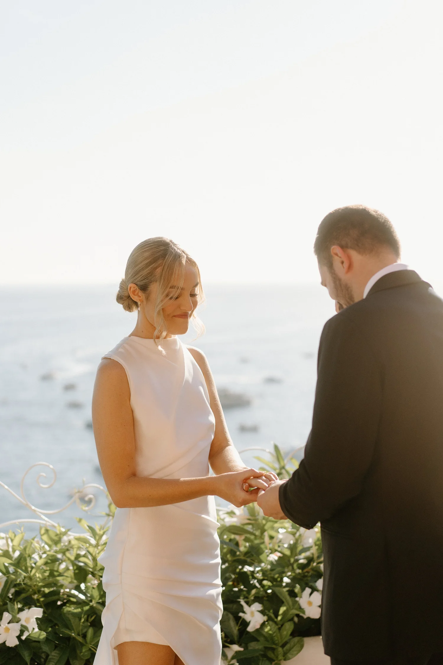A bride and groom holding hands during their outdoor wedding ceremony by the ocean, with greenery and white flowers in the background on a sunny day.