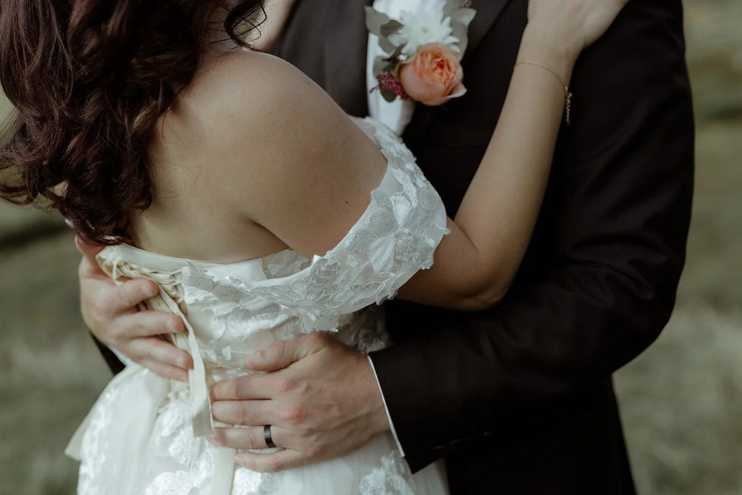 A close-up of a bride and groom embracing during their wedding. The bride is wearing an off-shoulder lace dress, and the groom is in a dark suit with a boutonniere featuring pink and white flowers.