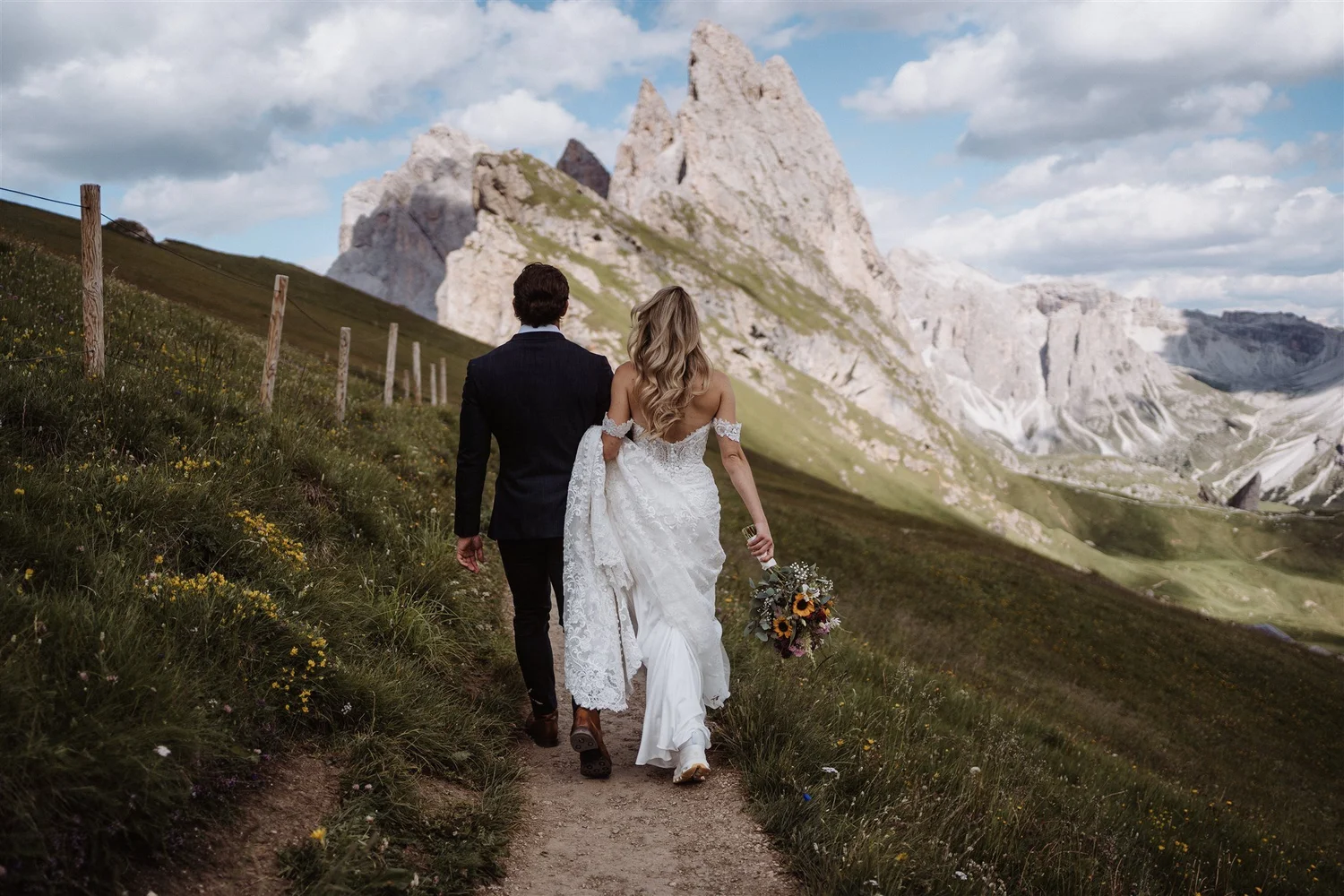 A newlywed couple walking on a trail in a mountainous landscape with rocky peaks, grass, and flowers under a partly cloudy sky.