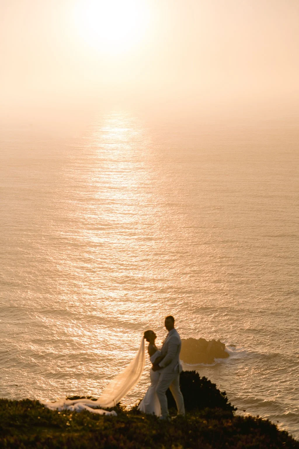 A couple in wedding attire stands on a grassy cliff by the ocean during sunset, with the bride's veil flowing in the breeze and the sun reflecting off the water.