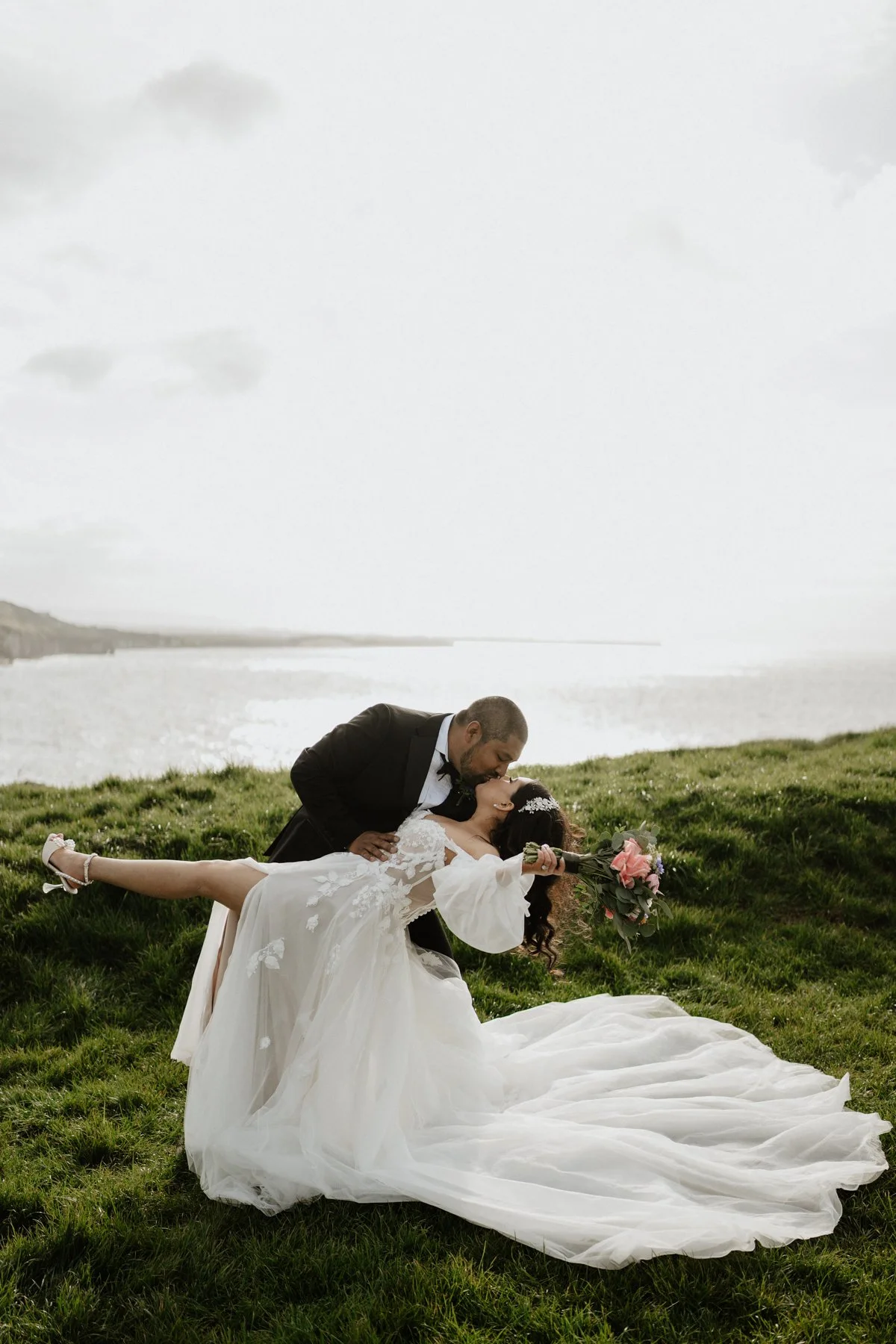 A groom in a black tuxedo and a bride in a white wedding dress are outdoors near a body of water. The groom is leaning over and kissing the bride, who is lying on the grass with her back arched and holding a bouquet of pink flowers. The bride's dress