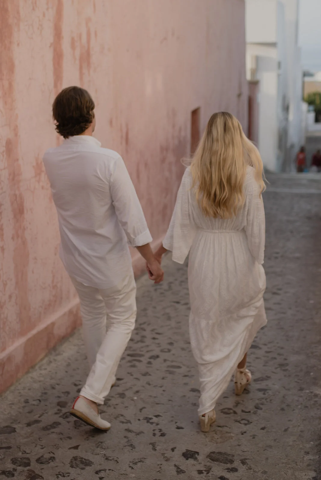 A couple walking hand in hand down a narrow cobblestone street, holding hands, with the woman in a long white dress and the man in white clothing.