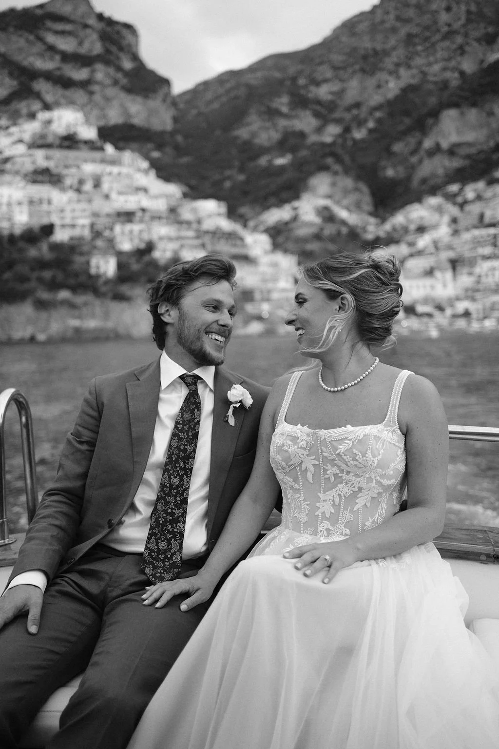 A black and white photo of a smiling couple dressed for a wedding, sitting close together on a boat with a mountain town in the background.