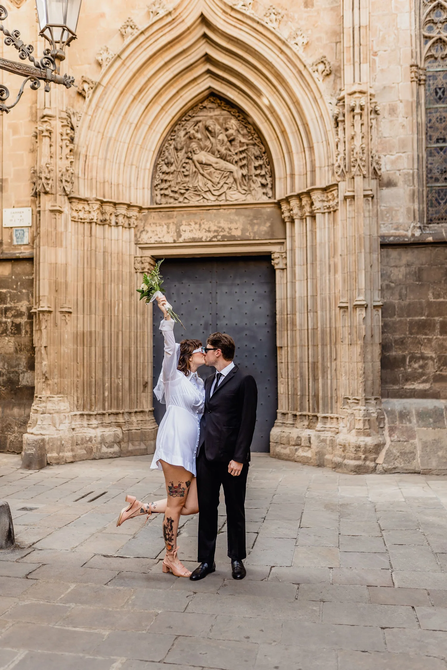 A couple dressed in wedding attire sharing a kiss in front of a historic stone building with ornate arches and carved sculptures. The woman is in a white dress, holding a bouquet, with tattoos on her legs, while the man is in a black suit.
