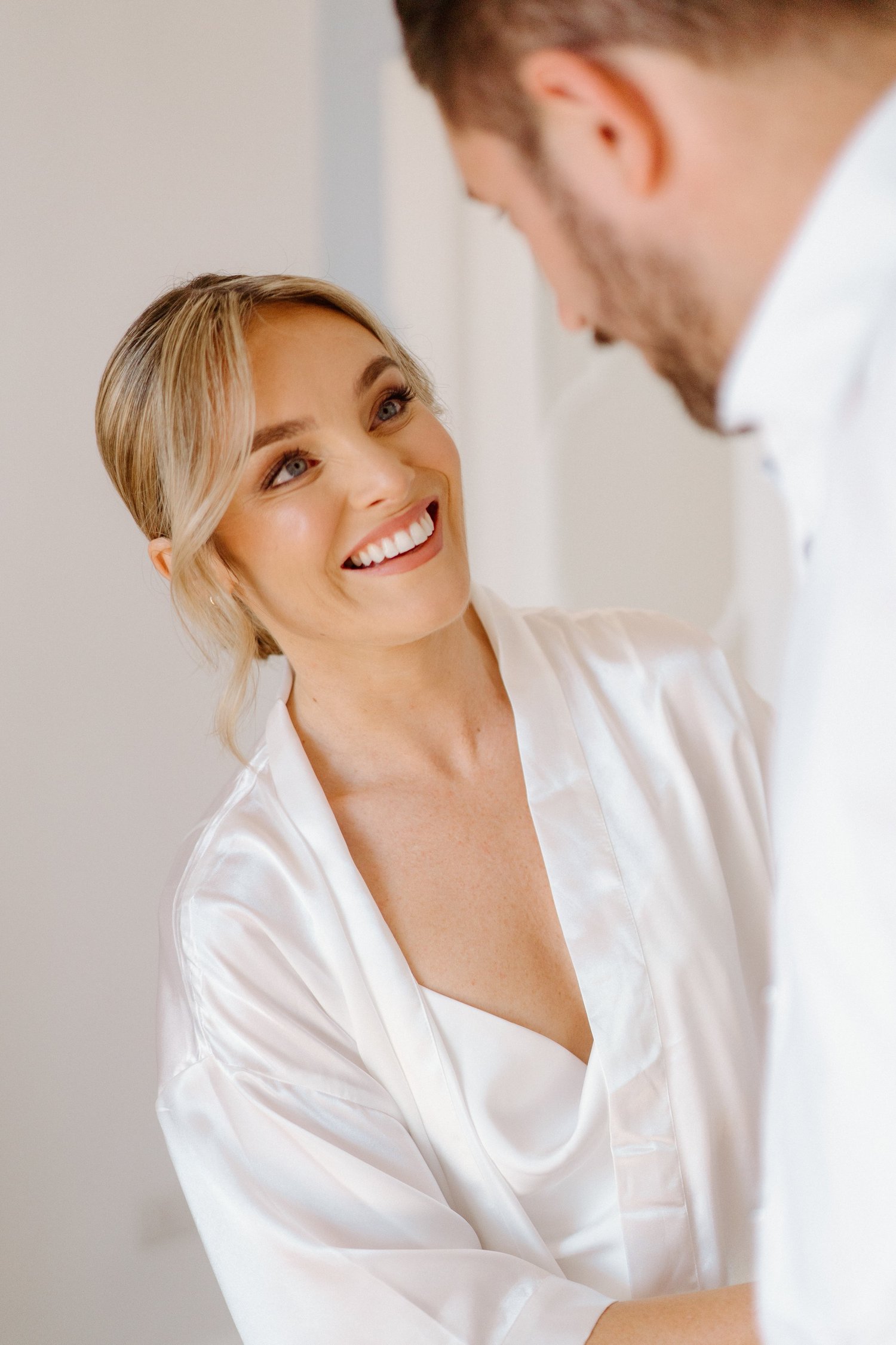 A woman with blonde hair styled in soft waves, wearing a white satin robe, smiling while looking at a man in a white shirt.