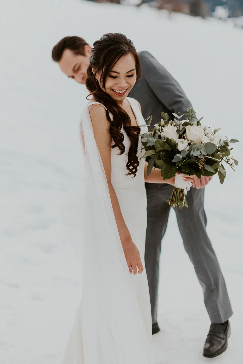 A happy bride with long dark hair in a white wedding dress, holding a bouquet of white roses and greenery.